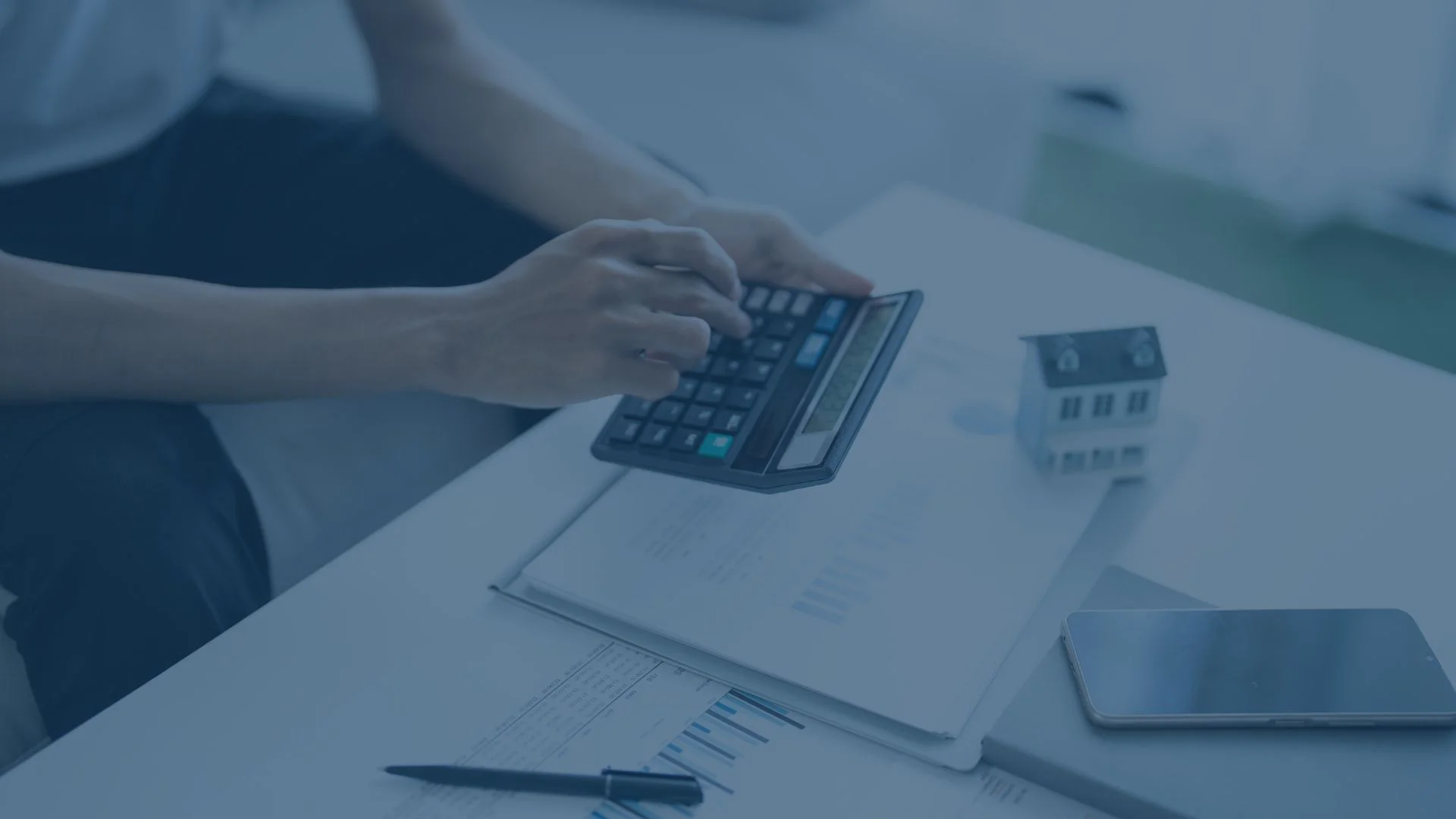 aksa.net Person berechnet finanzielle oder steuerliche Daten mit einem Taschenrechner, daneben liegen Dokumente und ein kleines Modellhaus.