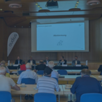 Teilnehmer einer Hauptversammlung sitzen vor einer Bühne mit Podium und Leinwand mit dem Titel „Abstimmung“