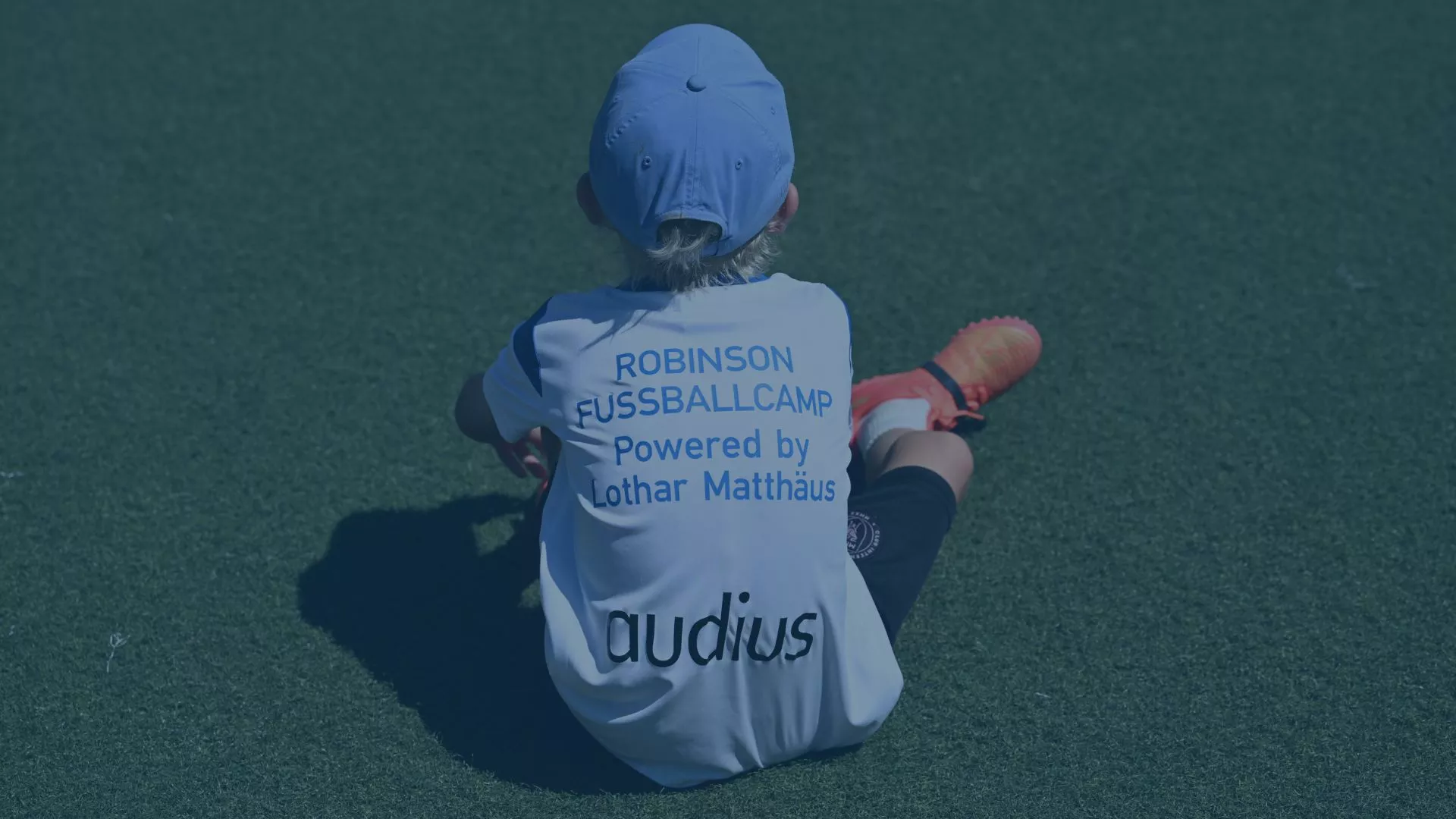 Ein Kind sitzt in Fußballkleidung auf einem Kunstrasenplatz, trägt ein Trikot mit der Aufschrift „Robinson Fußballcamp powered by Lothar Matthäus“ sowie dem Sponsorenlogo „audius“. Das Bild vermittelt die Idee der Nachwuchsförderung und des Teamgeists im Rahmen des Fußballcamps auf Kos, das jungen Sporttalenten sportliche Entwicklung und Begeisterung für Fußball ermöglicht.