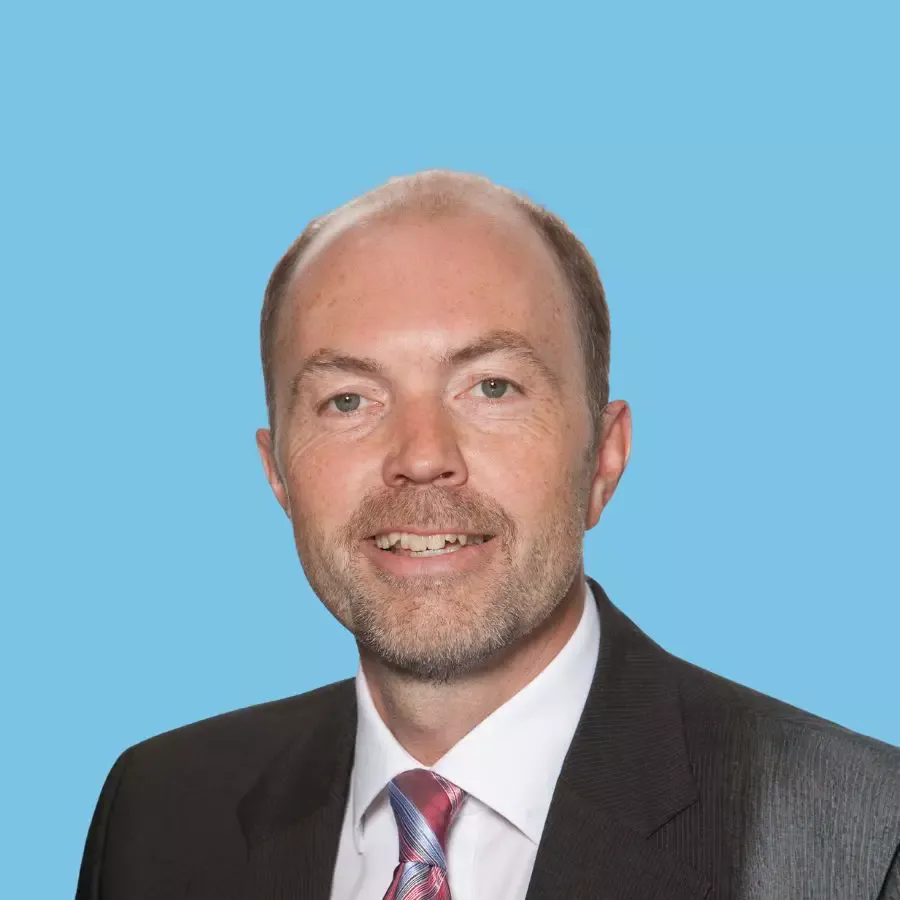 Porträt eines Mannes im Anzug vor hellblauem Hintergrund, der in die Kamera lächelt.