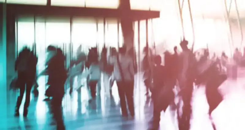 Verschwommenes Foto einer Menschenmenge, die durch einen hellen, modernen Raum mit Glaswänden und Sonnenlicht geht. Die Menschen sind in Bewegung und ihre Umrisse sind unscharf, was ein Gefühl von Hektik und Aktivität vermittelt.