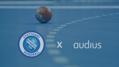 Das Bild zeigt ein Handball auf einem Hallenboden sowie die Logos des Handballvereins TVB 1898 Stuttgart und des Netzwerkpartners audius. audius unterstützt den TVB Stuttgart seit 2015